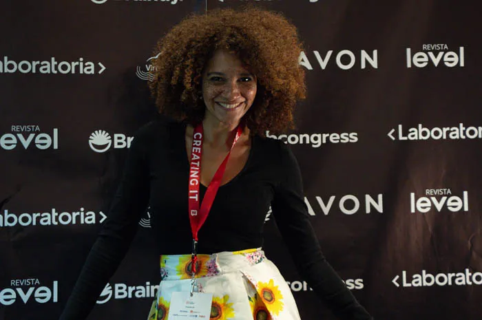 Mujer de cabello rizado sonríe frente al panel de patrocinadores de Revista Level y Avon en los Premios Mujeres Épicas 2022