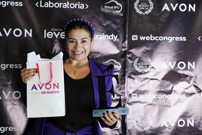 Mujer sonriente sostiene una bolsa de Avon y un trofeo en forma de letra frente al panel de Mujeres Épicas