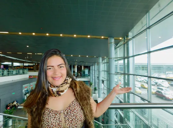 Retrato de una mujer sonriente con chaleco de leopardo y pañuelo, con un brazo extendido en un aeropuerto