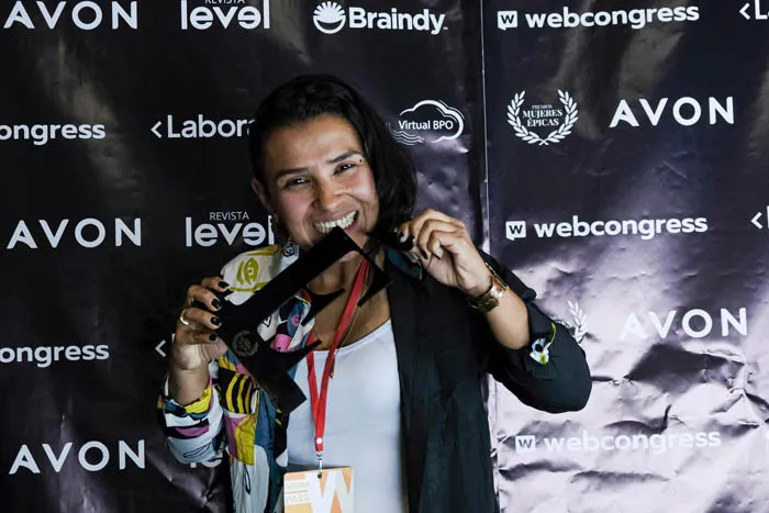 Mujer sonriente muerde su trofeo frente al panel de patrocinadores de Revista Level y Avon