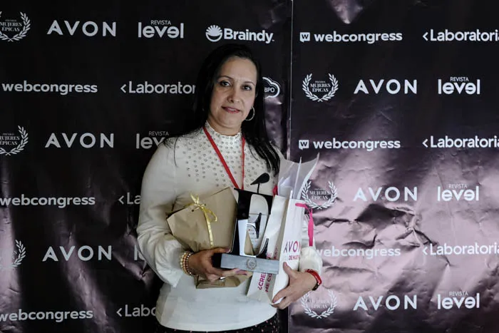 Mujer de suéter blanco sostiene un trofeo y un obsequio frente al panel de patrocinadores de Mujeres Épicas