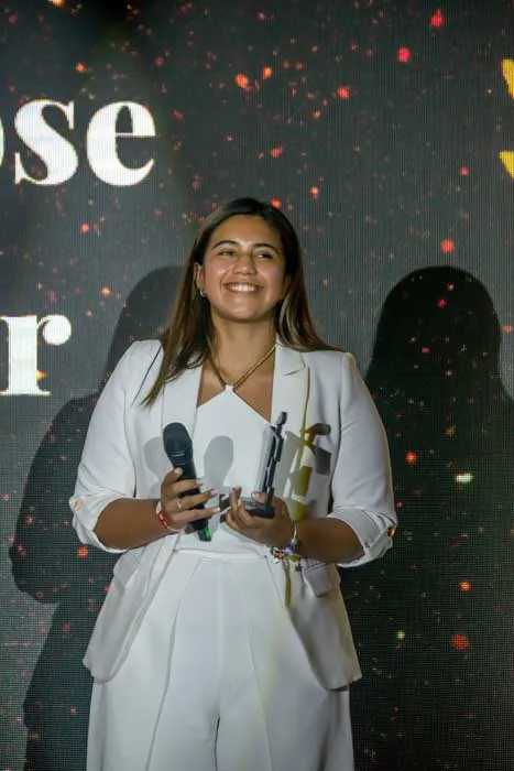 Mujer sonriente de traje blanco con micrófono y un trofeo en la mano sobre el escenario de Mujeres Épicas