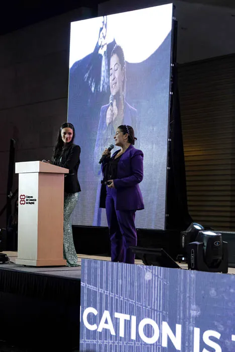 Mujer con traje morado habla en el escenario junto a un atril y una pantalla grande