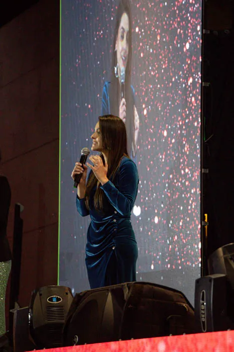 Mujer con vestido azul habla con un micrófono en el escenario ante una pantalla grande
