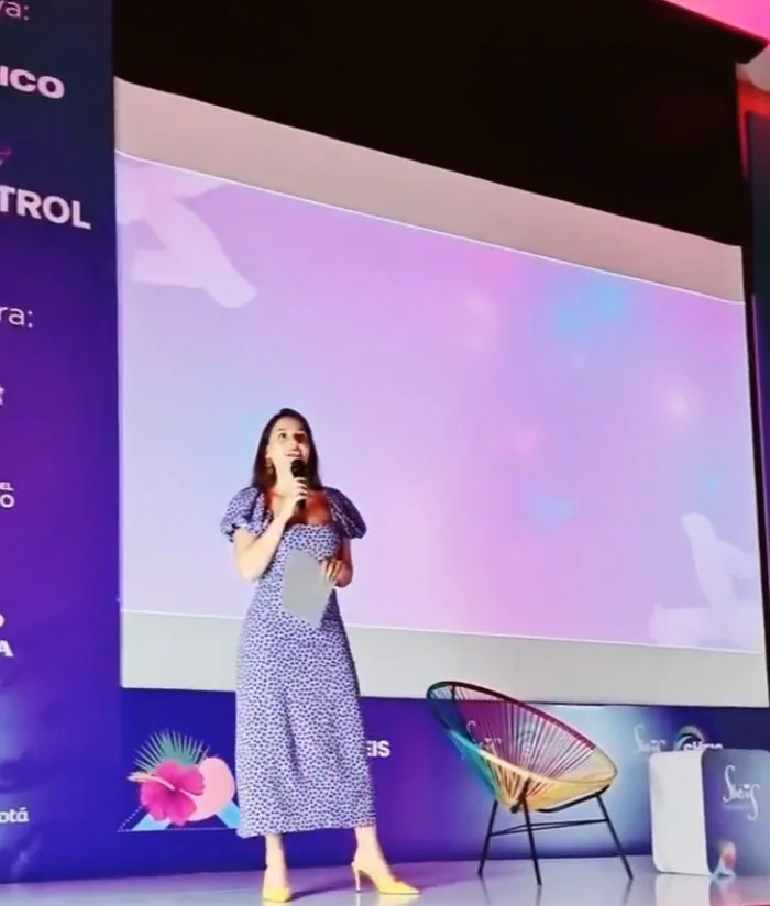 Mujer con vestido estampado y micrófono habla de pie ante una gran pantalla en el escenario del foro