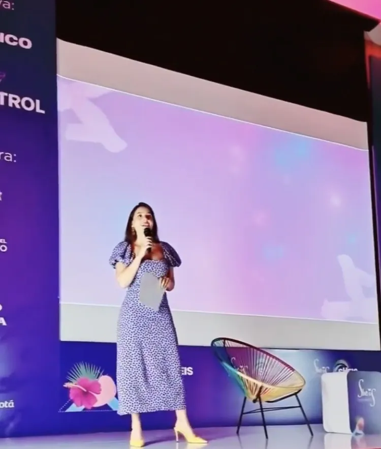 Mujer con vestido estampado y micrófono habla de pie ante una gran pantalla en el escenario del foro
