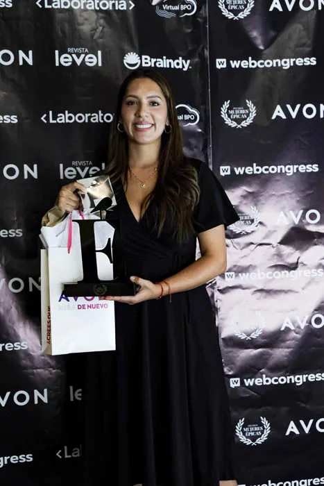 Mujer con vestido negro posa con un trofeo y una bolsa de regalo frente al panel de Revista Level y Avon