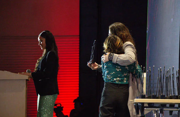 Dos mujeres se abrazan en el escenario junto a un atril y una mesa con trofeos