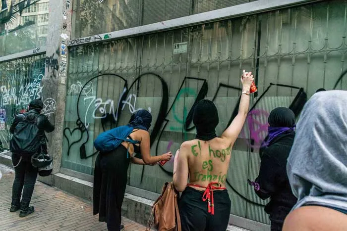 Mujeres pintan grafitis con aerosol sobre un muro de vidrio verde durante la marcha del 8 de marzo