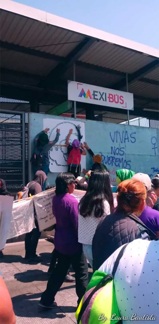 Mujeres con pañuelos y pancartas frente a una estación con la pinta «Vivas nos queremos» en la marcha del 8M