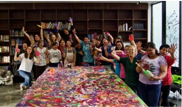 Grupo de mujeres con los brazos en alto tras una larga mesa cubierta de pintura de colores, en una biblioteca
