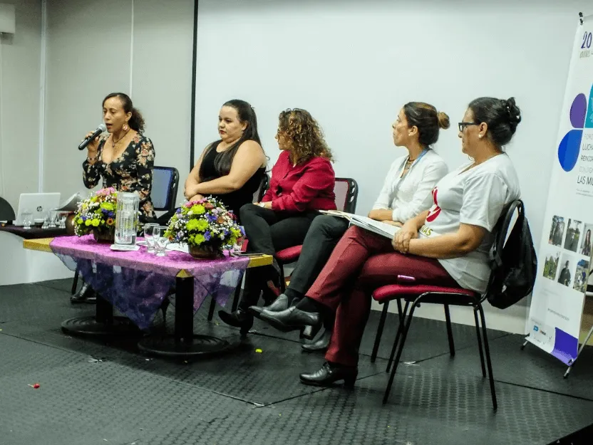 Cuatro mujeres sentadas en un panel detrás de una mesa con flores, una de ellas habla al micrófono
