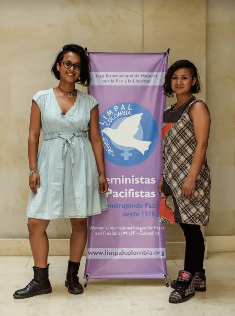 Dos mujeres posan junto a un pendón morado de «Feministas Pacifistas» con una paloma