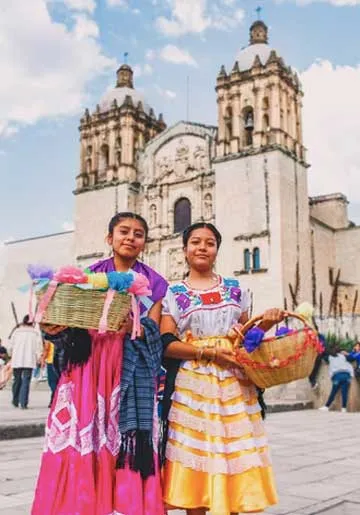 Dos mujeres con trajes tradicionales de colores vivos y canastas posan frente a una iglesia colonial de piedra