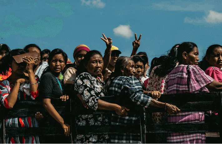 Multitud de mujeres con trajes tradicionales tras una valla bajo un cielo azul, algunas haciendo la señal de victoria