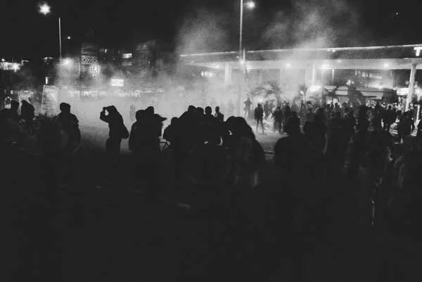Multitud en una calle de noche entre humo y luces durante una protesta, en blanco y negro