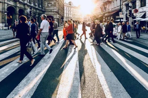 Multitud cruzando una calle en un paso de cebra al atardecer, con el sol al fondo