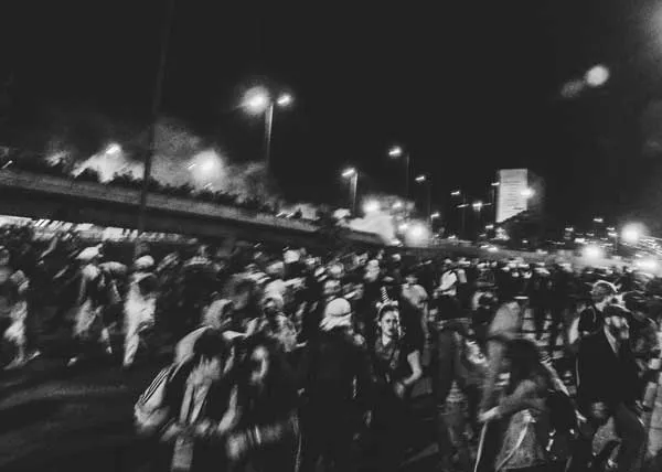 Multitud en movimiento junto a un puente bajo luces de calle de noche, en blanco y negro