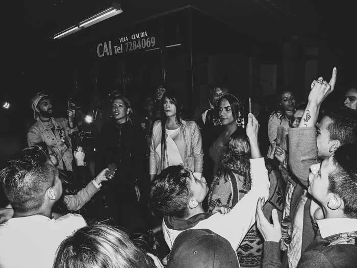 Multitud reunida de noche frente a un CAI de policía, con personas que levantan los brazos, en blanco y negro