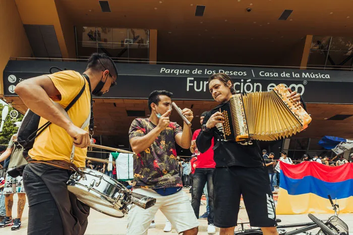 Músicos callejeros con acordeón y tambor tocan frente al Parque de los Deseos, junto a una bandera de Colombia