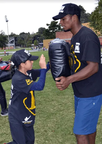 Niña con gorra golpea con la palma un escudo que sostiene un instructor en un parque