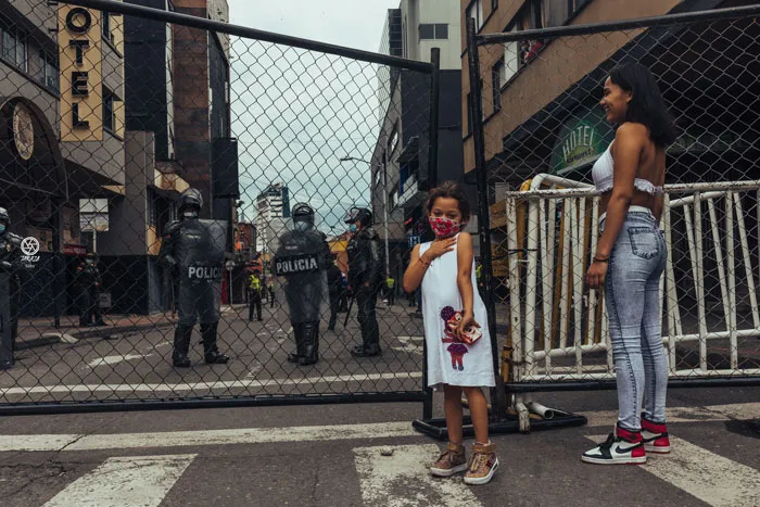 Una niña y una mujer de pie ante una reja metálica, con policías con escudos al otro lado de la calle