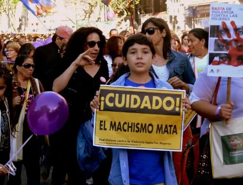 Niño en una marcha sosteniendo un cartel amarillo «¡Cuidado! El machismo mata» entre mujeres con globos morados