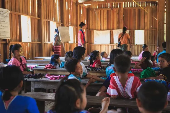 Niñas y niños sentados en pupitres de madera en un aula rural, con docentes al fondo