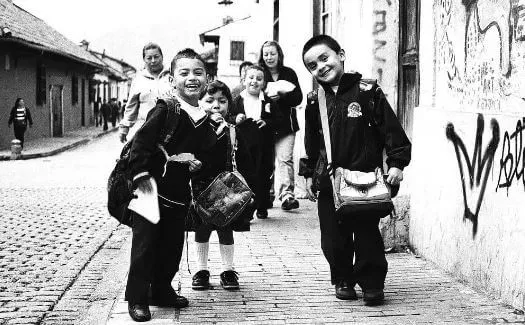 Niños con uniforme escolar sonríen mientras caminan por una calle empedrada con paredes grafiteadas