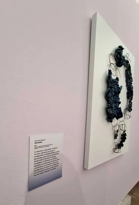 Obra de joyería azul oscuro con aros metálicos sobre un panel blanco, con su ficha explicativa