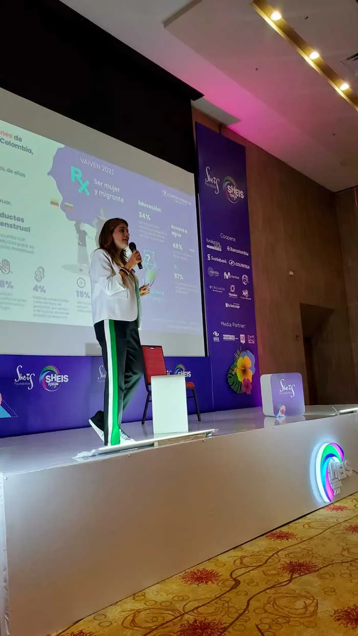 Oradora con pantalón deportivo de rayas habla de pie en el escenario del She is Global Forum ante una pantalla
