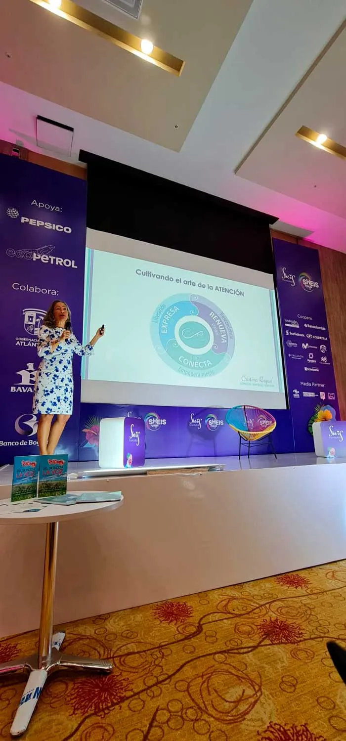Oradora con vestido estampado presenta en el escenario del She is Global Forum ante una diapositiva circular