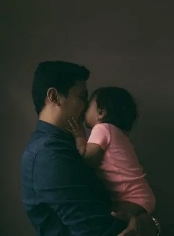 Padre cargando a su hija pequeña mientras la besa, en un ambiente oscuro