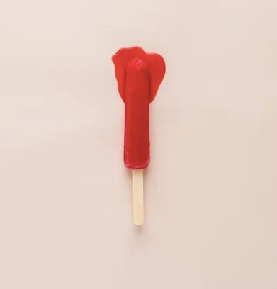Paleta de hielo roja derretida sobre un palo, con forma que evoca la vulva, sobre fondo rosa claro