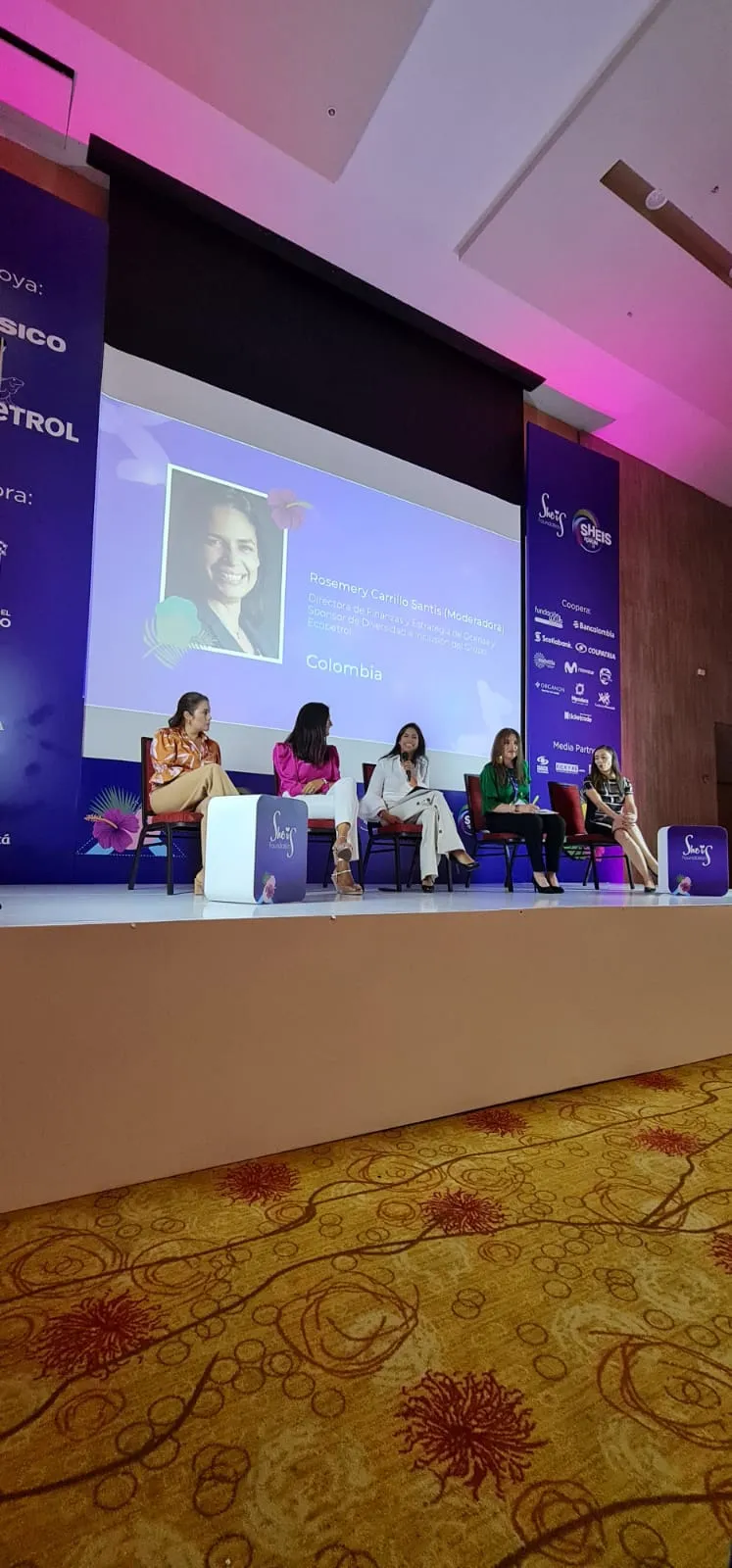 Panel de mujeres sentadas en el escenario con el retrato de una oradora proyectado en la pantalla