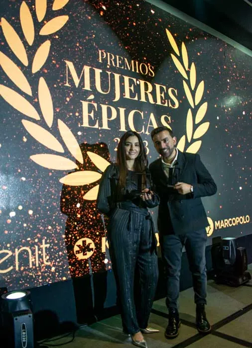 Una mujer y un hombre posan con sus reconocimientos frente al logo de los Premios Mujeres Épicas
