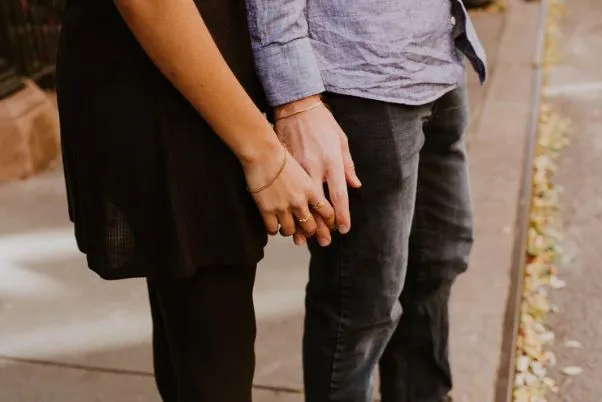 Pareja de pie tomada de la mano en una acera, vista de la cintura hacia abajo