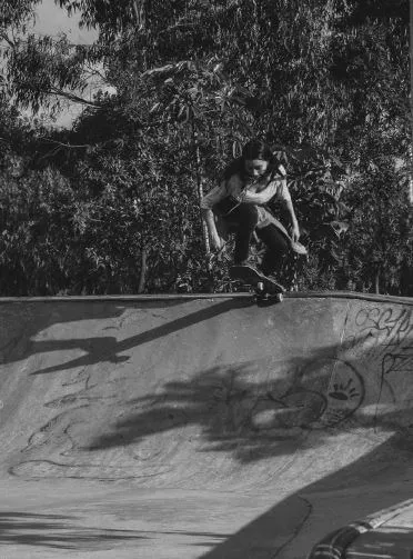 Patinadora en el aire sobre una rampa de skatepark bajo los árboles, en blanco y negro