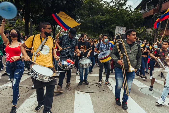 Percusionistas y un trombonista avanzan por un cruce peatonal entre banderas de Colombia