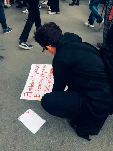 Persona vestida de negro en cuclillas escribe un cartel en el suelo durante una protesta en Ciudad de México