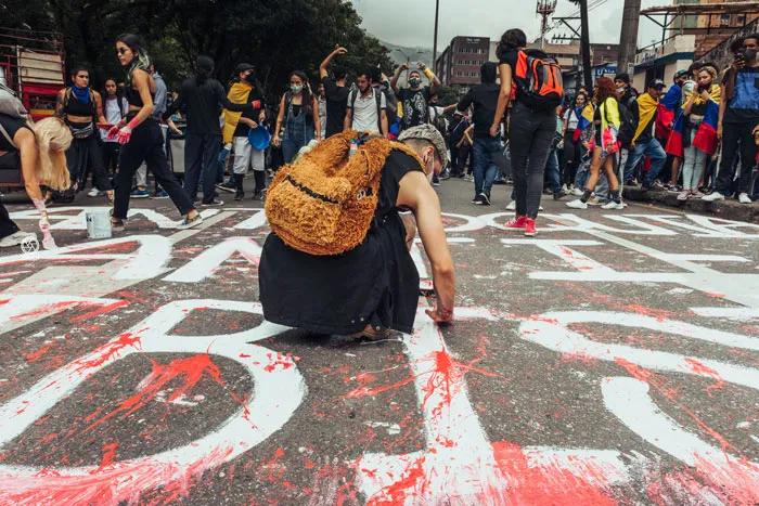 Persona agachada pinta letras rojas y blancas sobre el asfalto durante una manifestación