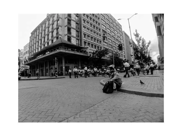 Persona sentada en el suelo de una plaza frente a un edificio grande, con una paloma cerca, en blanco y negro