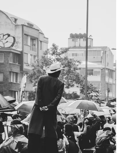 Persona con sombrero de espaldas frente a una multitud con paraguas y edificios grises, en blanco y negro