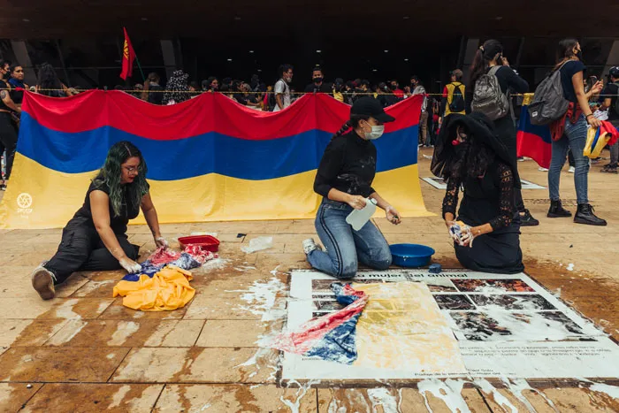 Tres personas de rodillas junto a una bandera de Colombia extendida y telas manchadas de pintura sobre el suelo