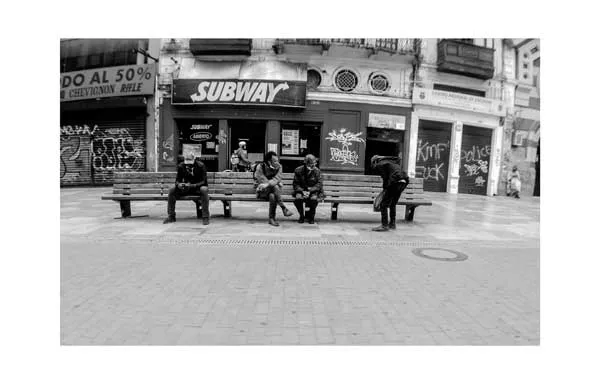 Personas sentadas separadas en una banca de una plaza casi vacía frente a un local, en blanco y negro