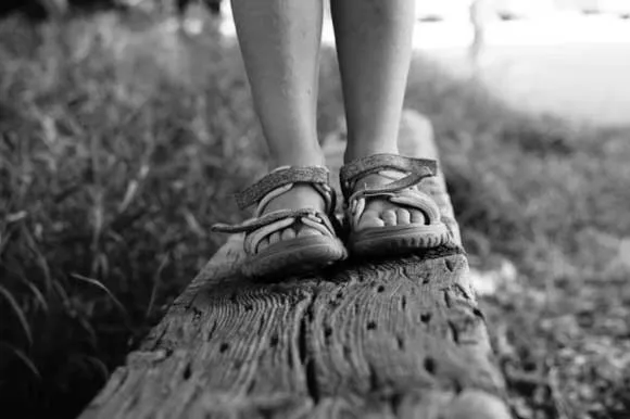 Pies con sandalias sobre un tronco de madera entre el pasto, en blanco y negro