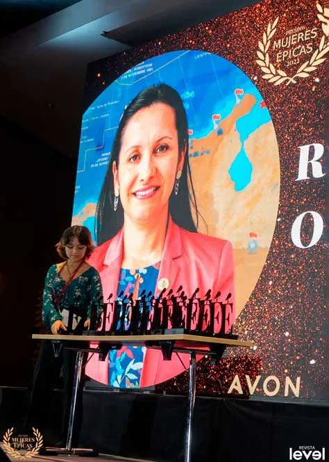 Presentadora junto a la mesa de premios frente a una pantalla con el retrato de una mujer, en Mujeres Épicas de Revista Level