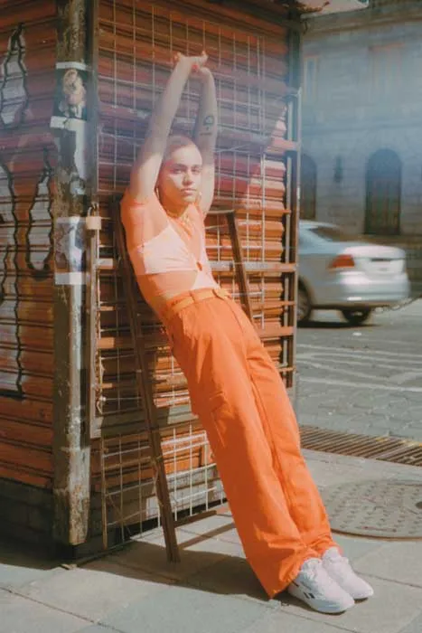Persona con conjunto naranja y zapatillas blancas, con los brazos en alto, apoyada en un quiosco en la calle