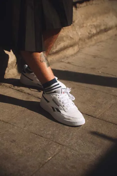 Piernas de una persona con falda negra, calcetines a rayas y zapatillas blancas Reebok sobre un pavimento
