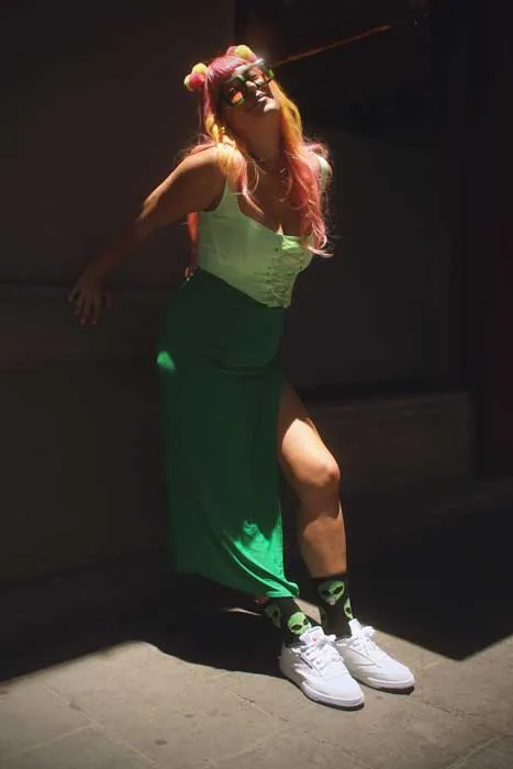 Persona de cabello rosado con falda verde larga y zapatillas blancas Reebok posa apoyada en una pared